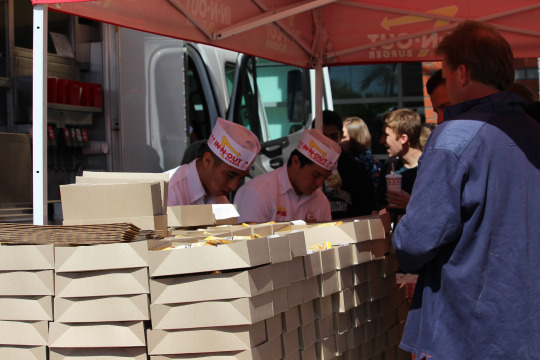 Two In-N-Out workers efficiently prepare lunches consisting of a signature burger, oven baked chips and a drink.