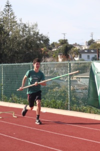 Freshman Aiden Parcell performs a  warm up while running with the pole vault stick and leaping over cones placed on the floor.